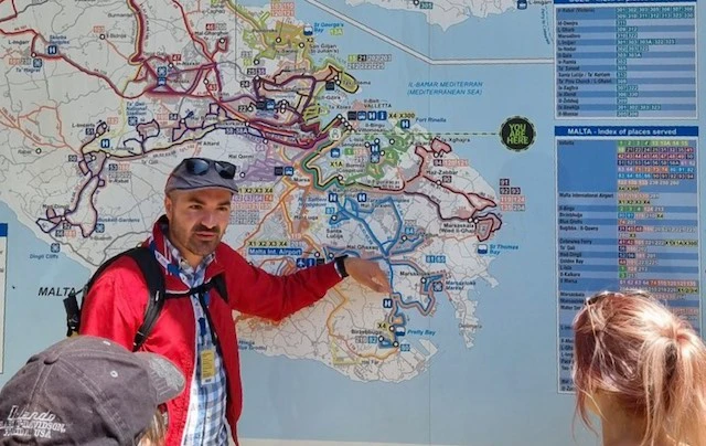 Picture of Ben pointing at a map of Malta with enraptured visitors watching